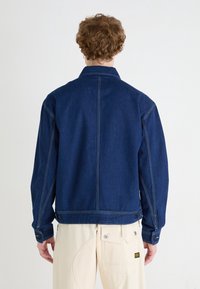 Persona con cabello rizado rojo, vistiendo una chaqueta de mezclilla azul oscuro y pantalones color crema, de espaldas contra un fondo blanco liso.