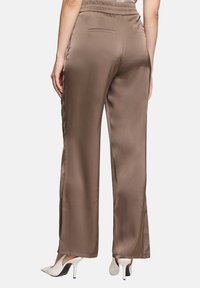 Braune Satin-Hosen mit weitem Bein und elastischem Bund, ausgestattet mit einer einzelnen Gesäßtasche und einer glatten Textur. Weiße High Heels sind sichtbar.