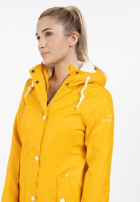 Kvinde med blond hestehale iført en lys gul hættetrøje med hvid foring og trykknapper, som står til venstre foran en hvid baggrund.