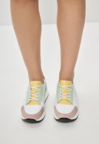 Zapatillas blancas con acentos en pastel amarillo y verde, detalles en suave gamuza rosa y cordones blancos planos. Diseño ligero y texturizado.
