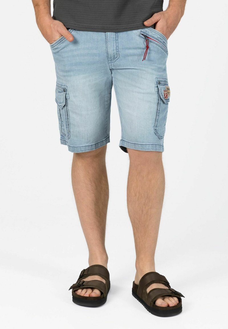 Helle blaue Denim-Shorts mit seitlichen Cargotaschen, einer vorderen Reißverschlusstasche mit roten Akzenten und einem locker sitzenden Schnitt. Getragen mit braunen Sandalen.
