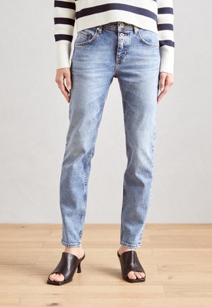 Personne portant un jean taille haute bleu clair et des mules noires à talons ouvertes, debout sur un sol en bois, avec un pull rayé blanc et marine.