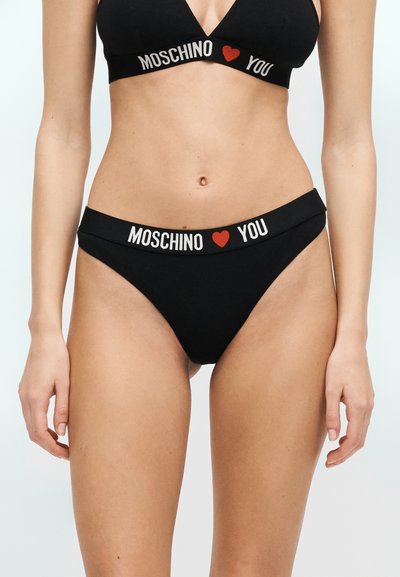 Femme portant un bikini noir de marque Moschino avec des symboles de cœur rouges entre "Moschino" et "You" sur les bandes, debout devant un fond uni.