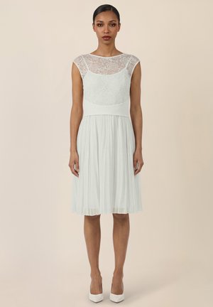 Vrouw draagt een knie-lange witte jurk met kanten lijfje en geplooide rok, staand tegen een effen beige achtergrond.