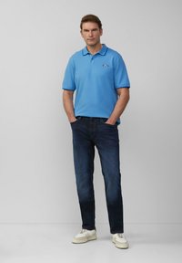 Man met een blauwe Ford polo, donkere jeans en witte sneakers, staand met handen in de zakken tegen een effen achtergrond.