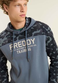 Felpa con corpo in tessuto blu, maniche a fantasia mimetica, cappuccio con coulisse e testo bianco con la scritta "FREDDY TRAINING."