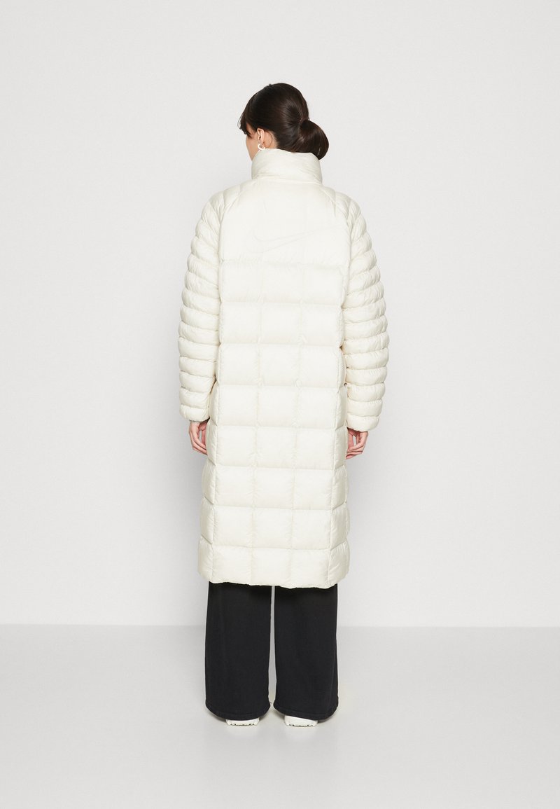 Manteau en duvet long de couleur beige clair avec un motif matelassé, col haut et manches élastiquées, porté sur un pantalon sombre. Son design minimaliste présente un logo.