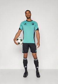 Tirkīza un melnas krāsas futbola tērps ar īsām piedurknēm, uz kura redzams Adidas logotips un trīs svītru akcenti. Modelis tur melni baltu futbola bumbu.