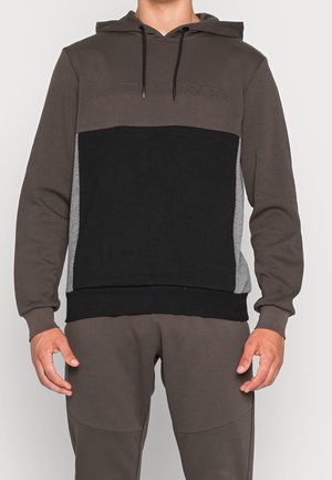 Man wearing a color-block hoodie with black front panel, brown sleeves and hood, and gray side panels, paired with brown pants.
