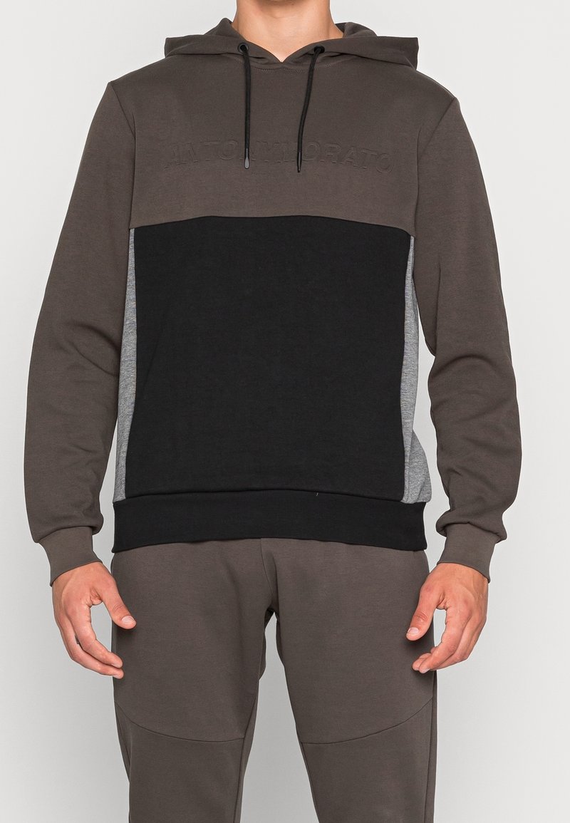 Homme portant un sweat à capuche color-block avec un panneau avant noir, des manches et une capuche marron, et des panneaux latéraux gris, associé à un pantalon marron.