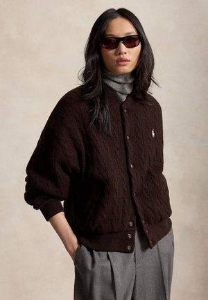 Donna che indossa un cardigan marrone scuro a trecce con bottoni, dolcevita grigio, pantaloni grigi e occhiali da sole scuri, in piedi contro uno sfondo beige.