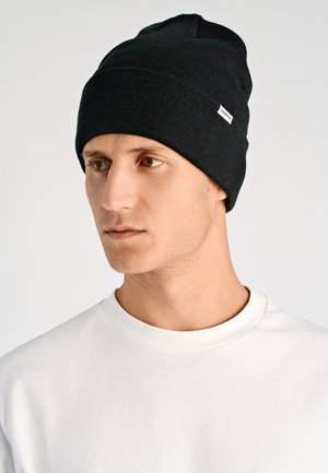 Giovane uomo che indossa un cappellino nero in maglia con un piccolo marchio e una maglietta bianca a girocollo, che guarda di lato su uno sfondo neutro.