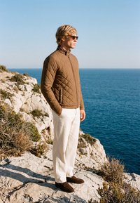 Uomo con giacca trapuntata marrone, pantaloni bianchi e occhiali da sole, in piedi su una scogliera rocciosa con vista sul mare azzurro calmo sotto un cielo sereno.