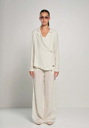 Vrouw draagt een loszittend, crèmekleurig linnen pak met lange mouwen en brede pijpen, staande tegen een effen lichte achtergrond.