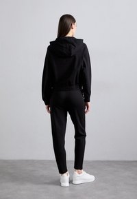 Moteris stovi atsisukusi atgal, vilkinti juodą trumpą gobtuvą, juodas sportines kelnes ir baltus sportbačius ant pilko grindų, priešais pilką sieną.