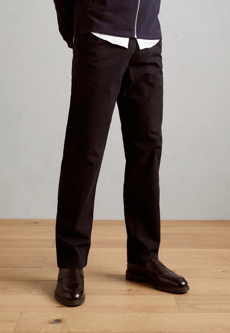 Pantalon en coton noir avec une coupe droite, associé à des chaussures en cuir marron foncé. L'arrière-plan présente un sol en bois clair.