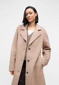 Παλτό από ανοιχτό καφέ μαλλί με γιακά τύπου notch, διαθέτει τέσσερα κουμπιά και δύο τσέπες. Υφή υφάσματος με ανάγλυφο μοτίβο και χαλαρή σιλουέτα.