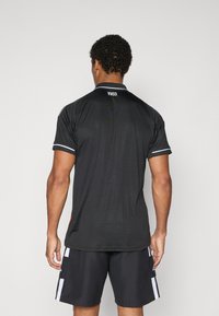 Juoda sportinė polo marškinėliai su apykakle, trumpomis rankovėmis ir kontrastingu baltu apvadu. Gale yra mažas etikietė su užrašu „1903“.