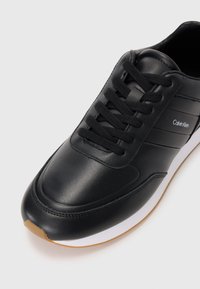 Crni sintetički tenisice s glatkom teksturom, sadrže šest crnih vezica, bijelu srednju potplat i smeđi gumenjak. Vidljiv Calvin Klein logo.