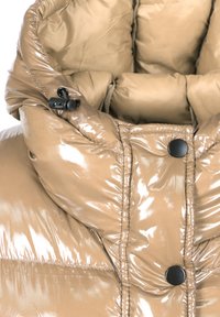 Glanzende beige pufferjack met zwarte drukknoopjes en verstelbare koordversteller op de hoge kraag voor extra warmte.