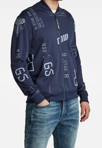Chaqueta navy con cremallera y texto y gráficos impresos en blanco. Cuenta con un cuello y puños acanalados. Combinada con jeans de color azul claro.