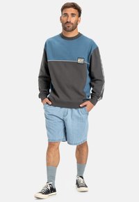 Felpa color block grigia e blu con polsini a coste, patch del logo sul petto, abbinata a pantaloni corti di jeans chiari e sneaker nere.