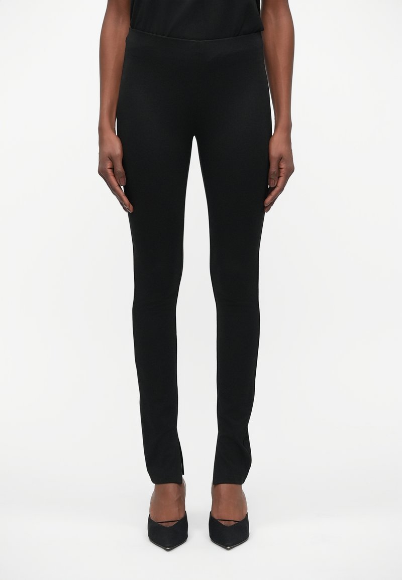 Zwarte leggings gemaakt van rekbare, gladde stof. Strakke pasvorm met een slanke vorm en subtiele zijsplitsingen aan de zoom. Gekleed met zwarte hakken.