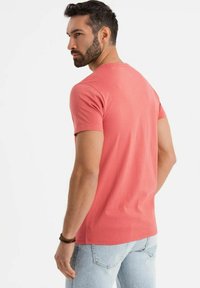 Korálové bavlněné tričko s klasickým střihem a krátkým rukávem, viděné zezadu. Světle modré džíny zdůrazňují vzhled.