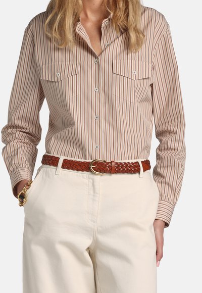 Ženska, oblečena v bež in rjavo črtasto srajco na gumbe, zataknjeno v kremne visoke hlače z rjavo usnjeno pleteno pasom.