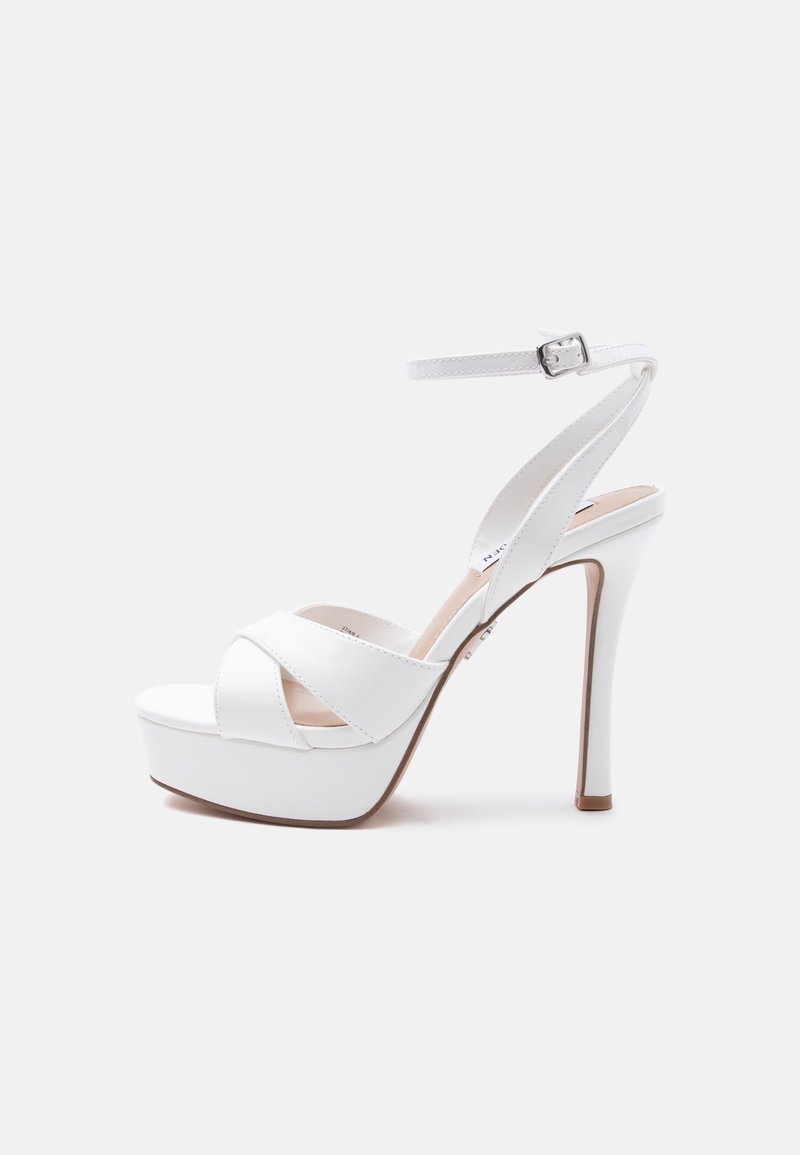 Λευκό πέδιλο πλατφόρμας με ψηλό τακούνι, λουράκι στον αστράγαλο με αγκράφα, ανοιχτά δάχτυλα και λουράκια που διασταυρώνονται μπροστά.