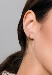 Złote kolczyki w kształcie obręczy z mniejszym okrągłym wzorem, akcentem w postaci kryształu, teksturowaną powierzchnią i gładkim wykończeniem, noszone na uchu modelki.