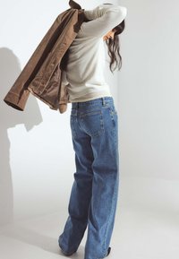 Brūnas zamša jaka, balts garas piedurknes tops un zili plaša kāju džinsi. Džinsi ir brīvā piegriezumā ar klasiskām aizmugurējām kabatām un šuvju detaļām.