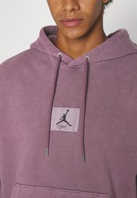 Sudadera púrpura hecha de tela suave con un bolsillo tipo canguro, que presenta una etiqueta con un logo negro de Jordan y cordones con puntas de metal.