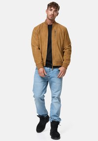 Brown suede bomber jacket with a front zipper and side pockets, worn over a black shirt, paired with light blue jeans and black boots.