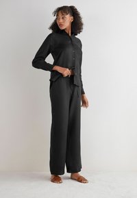 Svart skjorta med knappar, lång ärm och ficka, i kombination med svarta lösa byxor. Bärs med bruna sandaletter på en ljus yta.