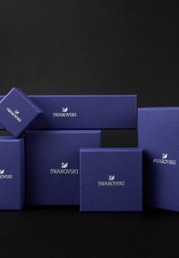 Sammlung von lila strukturierten Boxen mit weißem "Swarovski"-Logo, die in verschiedenen Größen und in unterschiedlichen Winkeln auf einem schwarzen Hintergrund angeordnet sind.