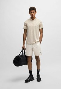 Camisa polo beige con un patrón sutil y el logo de BOSS, combinada con pantalones cortos color crema. Una bolsa deportiva negra, calcetines negros y zapatillas oscuras completan el look.