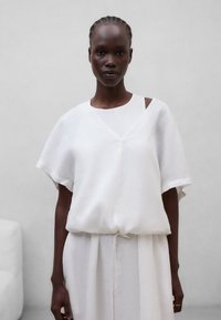 Personne à la peau foncée avec des cheveux tressés portant un haut ample blanc à manches courtes sur une robe blanche assortie, debout devant un fond gris clair uni.