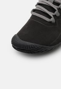 Scarpa sportiva nera con suola in gomma texture, parte superiore liscia e lacci grigi a contrasto. Presenta un design con punta arrotondata.