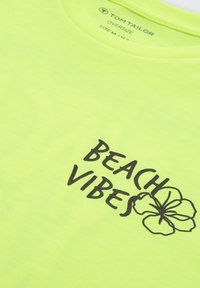 Neon dzeltas liela izmēra T-kreklu ar apaļu kakla izgriezumu, kas apdrukāts ar melnu tekstu "BEACH VIBES" un ziedu dizainu. Mīksta tekstūra, ikdienišķs dizains.