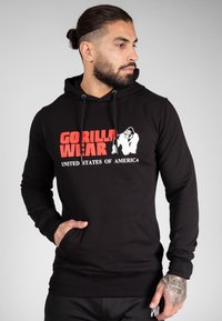Hoodie noir avec cordons de serrage, affichant un audacieux graphique rouge et blanc "GORILLA WEAR" et "ÉTATS-UNIS D'AMÉRIQUE" à l'avant. Texture lisse.