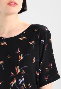 Blusa negra con un patrón de aves coloridas, mangas cortas y un escote amplio, hecha de una tela ligera y texturizada.