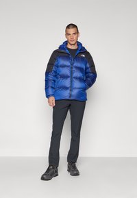 Niebieska kurtka puchowa z czarnymi ramionami, zapięcie na zamek i kaptur. Noszona z ciemnymi spodniami i czarnymi butami. Gładka, błyszcząca tekstura.