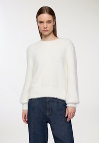 Kobieta z prostymi brązowymi włosami nosząca biały puszysty sweter i ciemnoniebieskie dżinsy z wysokim stanem, na jednolitym jasnym tle.