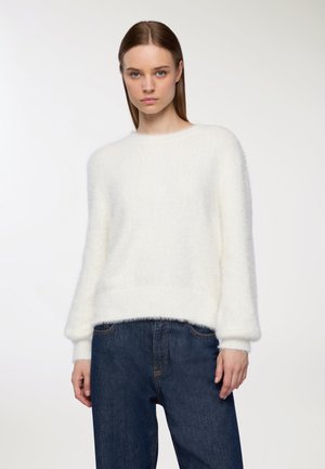 Donna con capelli castano lisci che indossa un maglione bianco morbido e jeans a vita alta blu scuro, su uno sfondo chiaro e semplice.