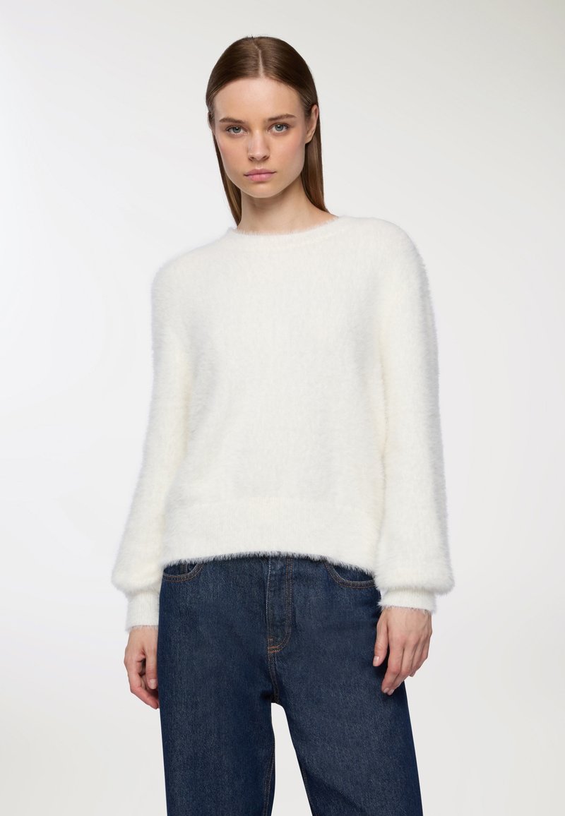 Kobieta z prostymi brązowymi włosami nosząca biały puszysty sweter i ciemnoniebieskie dżinsy z wysokim stanem, na jednolitym jasnym tle.