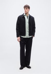 Νεαρός άνδρας στέκεται φορώντας μαύρο σακάκι πάνω από ανοιχτοπράσινο πουκάμισο με κουμπιά, μαύρο παντελόνι και μαύρα παπούτσια, με τα χέρια στις τσέπες σε απλό φόντο.