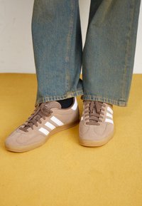 Sneakers in camoscio marrone con strisce bianche, punta tonda e design con lacci. Suola in gomma e dettagli in rilievo. Indossate con jeans di denim blu.