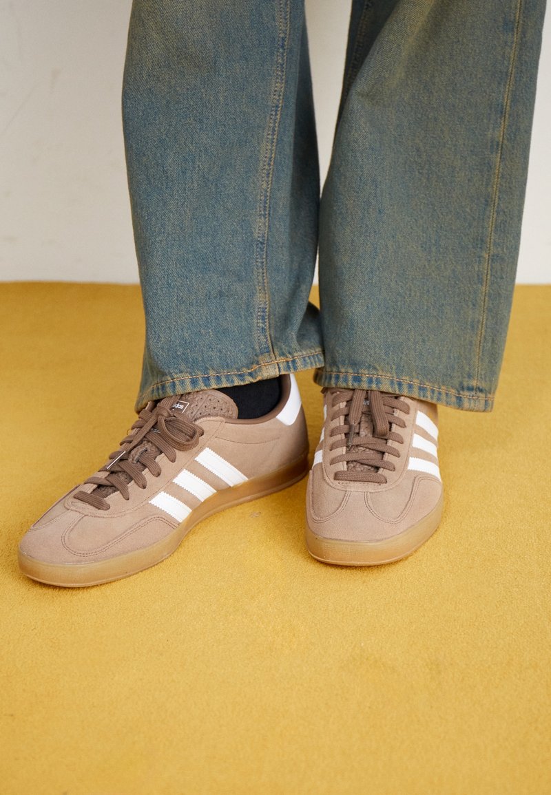 Sneakers in camoscio marrone con strisce bianche, punta tonda e design con lacci. Suola in gomma e dettagli in rilievo. Indossate con jeans di denim blu.