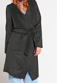 Ciemnoszara wełniana płaszcz o kroju narzutki z dekoltem w kształcie V, szerokimi klapami i paskiem do wiązania. Posiada przednią kieszeń oraz asymetryczny dół.